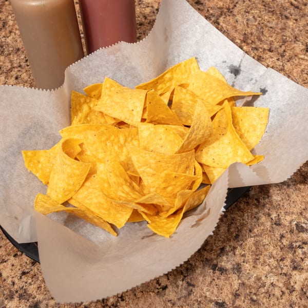 A basket of crispy tortilla chips with two sauce bottles in the background on a speckled counter.