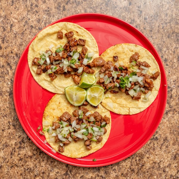 Three street tacos with diced meat, onions, cilantro, and lime wedges on a red plate.