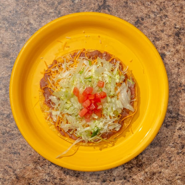A tostada with refried beans, shredded cheese, lettuce, and diced tomatoes on a yellow plate.