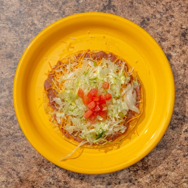 A tostada with refried beans, shredded cheese, lettuce, and diced tomatoes on a yellow plate.